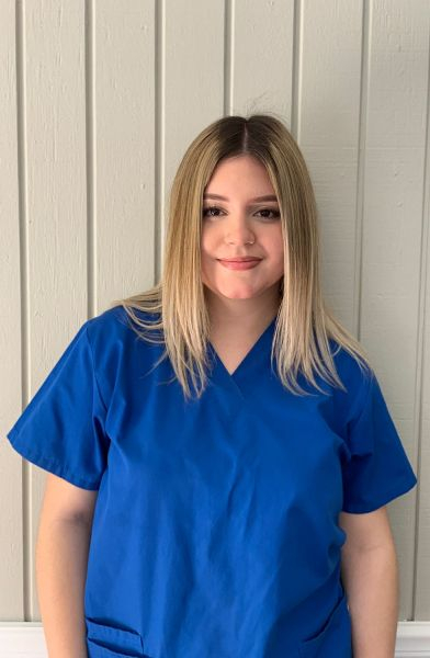 Young woman with blonde hair wearing a blue scrub top, smiling gently.