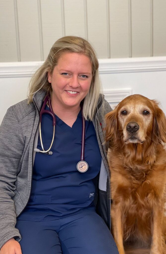 A smiling woman in medical scrubs seated next to a golden retriever.
