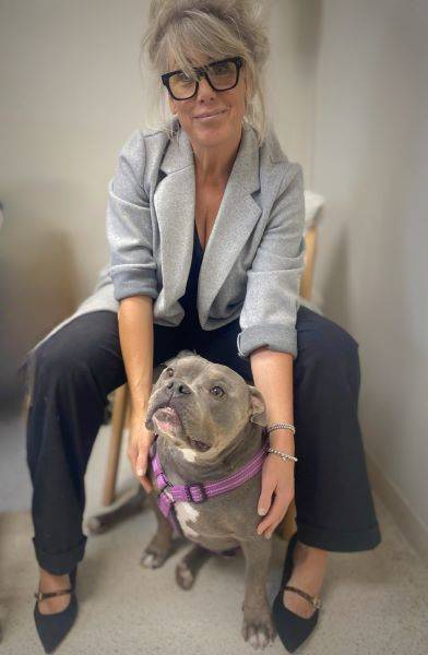 Woman in a gray blazer and glasses sitting with a gray pit bull wearing a pink collar.