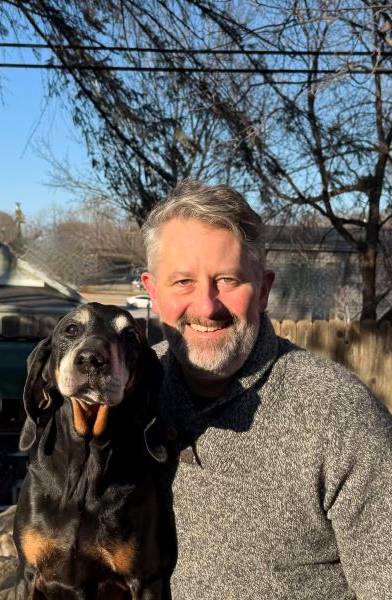Man with a beard and a black dog smiling outdoors in sunlight.